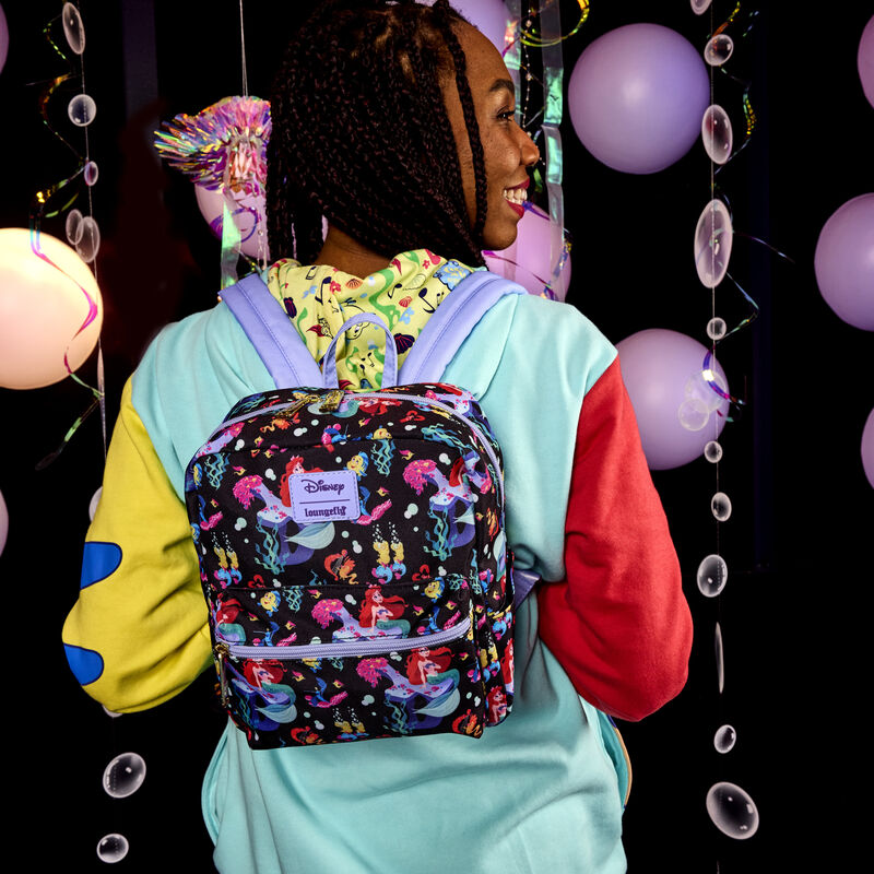 Woman facing away from camera wearing the Loungefly Little Mermaid 35th Anniversary Ariel & Flounder Glow Unisex Hoodie and the Little Mermaid 35th Anniversary Life is the Bubbles All-Over Print Nylon Square Mini Backpack, featuring a repeating pattern of Ariel, Sebastian, Flounder, and other aquatic musicians and motifs. 